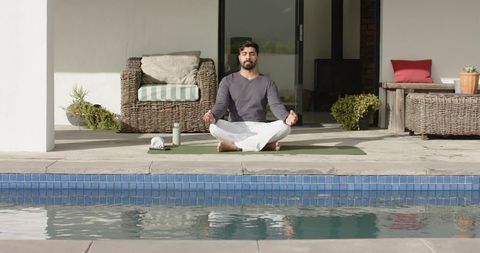 Indian man meditating by pool on patio yoga mat amid wicker furniture and plants