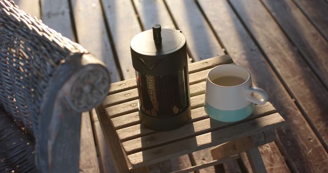 French press and ceramic coffee cup sitting on wooden table bathed in warm morning sunlight. Ideal for illustrating relaxing morning routines, promoting coffee-related products, or depicting serene outdoor scenes.