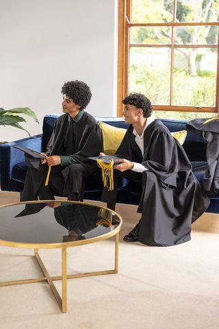 African american male graduates sitting on velvet sofa at home holding gold-tassel caps