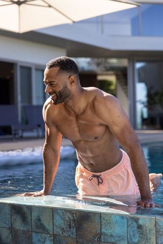 African mature man emerging from pool on modern backyard patio wearing pink swim trunks