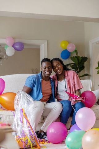 African american couple laughing and sharing gift in pastel balloon-decorated living room