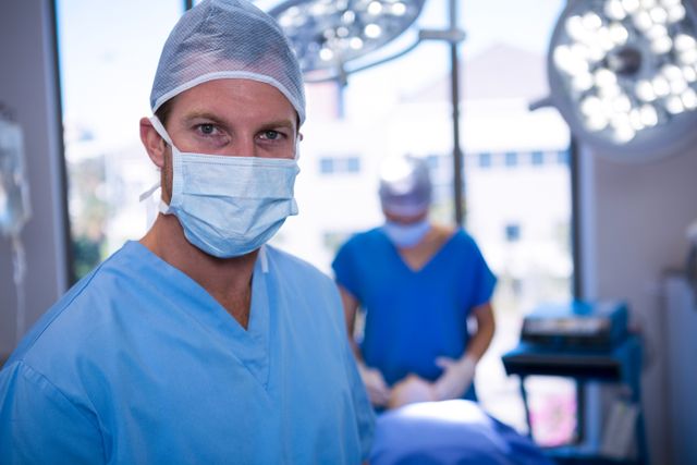 Male nurse wearing surgical mask in operating room, standing in foreground while another medical professional works in the background. Ideal for use in healthcare, medical, and hospital-related content, showcasing professionalism and sterile environment in surgical settings.
