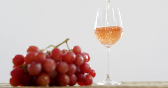 Rosé wine is being poured into a clear glass beside a bunch of fresh, red grapes on a light wooden surface. This image can be used for websites, articles or marketing materials related to wine, beverages, fine dining, and culinary experiences. Also suitable for advertisements and packaging designs in the wine and beverage industry.