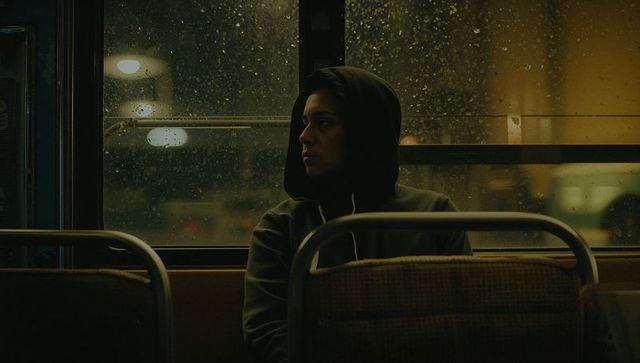 Solitary woman in hoodie gazing out rain-speckled bus window at night, urban transit mood