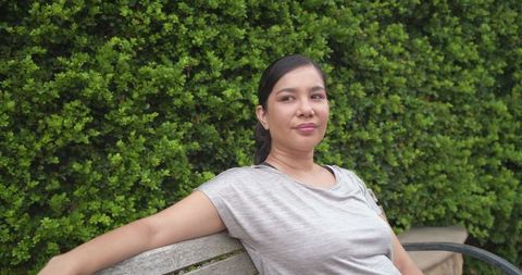 Relaxing mid-adult woman reclining on bench beside lush hedge enjoying calm outdoors
