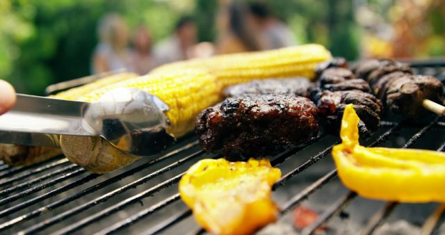Corn on the cob and meat kebabs are grilling over an outdoor barbecue grill. Tongs are being used to turn a piece of meat. The blurred background suggests a lively gathering, ideal for holdings event promotions, barbecue-themed advertisements, food blogging visual content, or seasonal campaigns celebrating outdoor dining and summer activities.