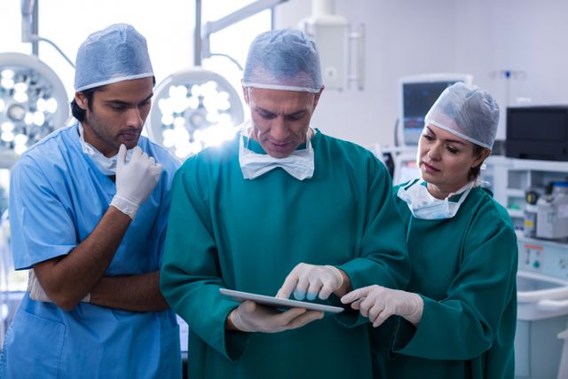 Team of surgeons in surgical attire discussing over a digital tablet in an operating room. Ideal for use in healthcare, medical technology, and teamwork-related content. Suitable for illustrating modern medical practices, surgical procedures, and professional collaboration in hospitals.