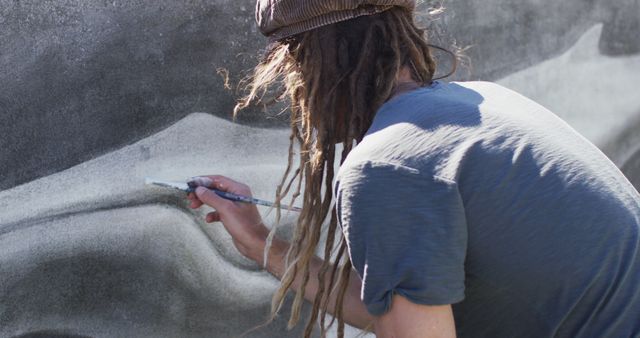 Street artist with dreadlocks creating large mural on outdoor wall with broad brushstrokes. Suitable for use in articles about urban art, street culture, creativity, or mural projects.