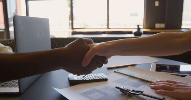 Two people are shaking hands across table with laptops and papers. Sunlight filters through large windows, highlighting their professional handshake