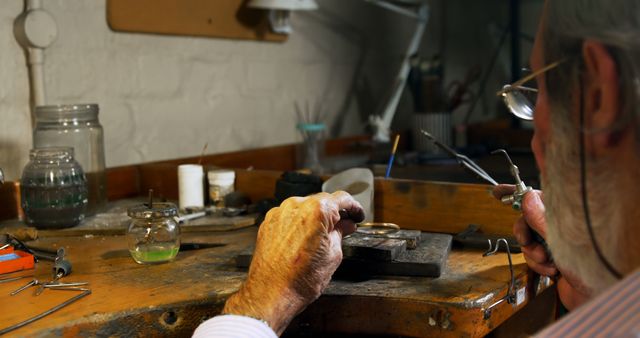 Elderly craftsman meticulously creating a piece of jewelry in his workshop. Surrounded by essential tools and equipment, he employs skilled techniques to shape and refine. Perfect for illustrating themes of craftsmanship, skilled trades, dedication, and the art of handmade objects. Can be used in articles about career craftsmanship, jewelry making, and workshop settings.