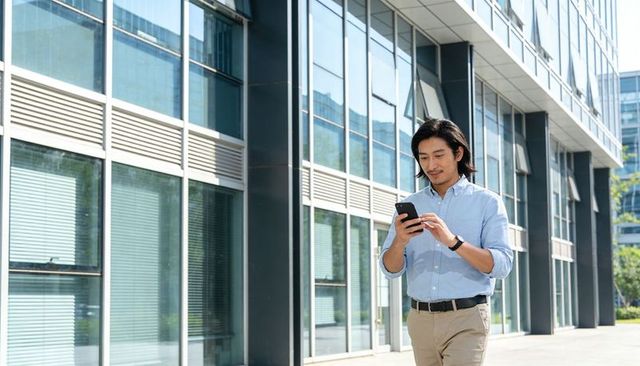 Young professional walking and checking smartphone beside urban modern glass office facade