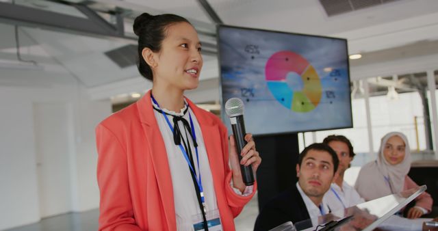 Side view close up of a young Asian businesswoman wearing a pink jacket standing at a lectern using a microphone to address the audience at a business conference. In the background is a screen with information on it and a diverse group of business people sitting, looking up at her and listening
