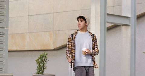 Asian man traveling with carry-on and coffee standing in modern urban plaza