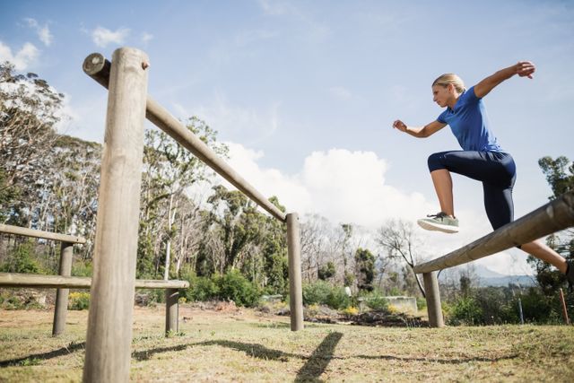 Woman participating in an outdoor obstacle course, jumping over wooden hurdles. Ideal for fitness, training, and healthy lifestyle themes. Can be used in advertisements for boot camps, athletic training programs, or motivational content.