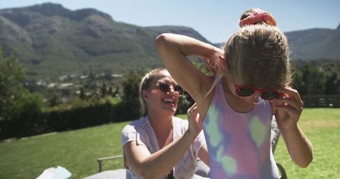 Mother laughing with daughter adjusting red sunglasses in pastel swimsuit on sunny lawn