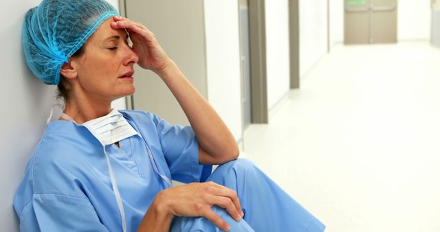 Female nurse in blue scrubs sitting against hospital corridor wall, touching forehead, showing stress and fatigue. Useful for depicting healthcare industry struggles, stress management, healthcare worker appreciation, mental health, and the challenges faced by medical professionals.
