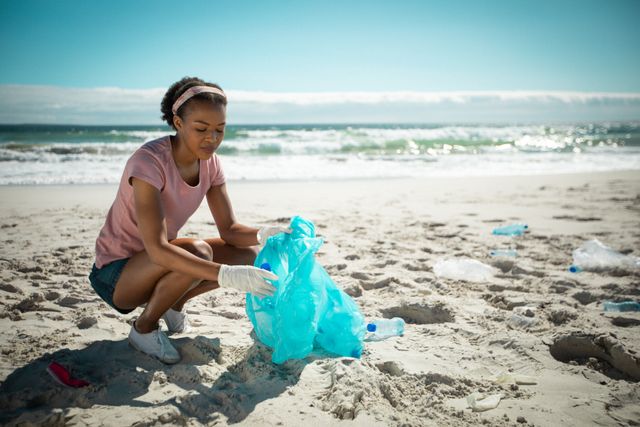 African american woman putting rubbish in refuse sack on sunny beach. eco conservation volunteer, beach clean-up.