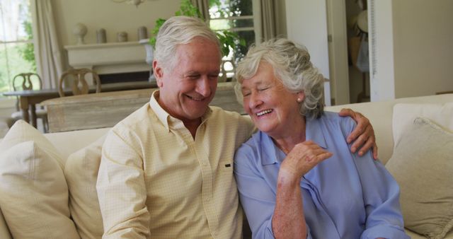 A senior couple sitting together on a sofa, joyfully smiling and embracing at home. This cheerful moment highlights their strong relationship and happy retirement life. Ideal for use in advertisements or promotions related to senior living, retirement communities, healthcare services for the elderly, family life, or any content celebrating love and relationships in later years.