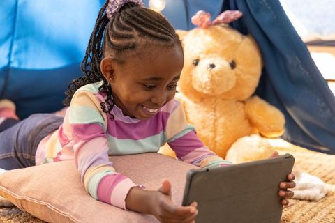 Smiling african american girl using tablet inside cozy indoor tent with giant teddy bear