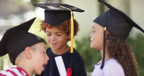 Diverse children celebrating graduation wearing mortarboards and holding diplomas outdoors