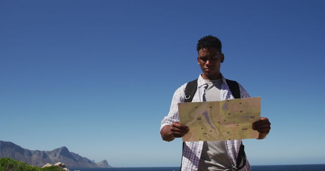 Young man reading map during mountain hike under clear blue sky, holding map, backpack on shoulders. Ideal for content about travel, hiking adventures, nature exploration, navigation skills, and outdoor activities.