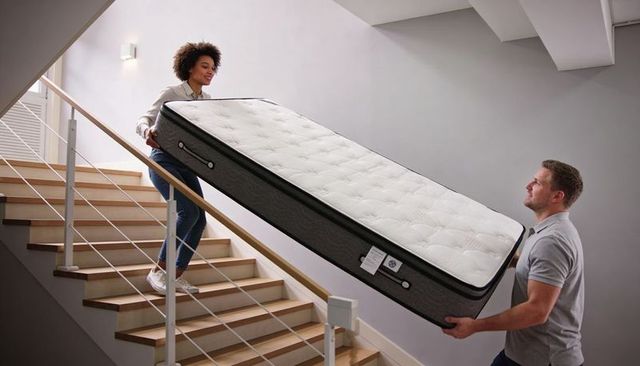 Couple carrying queen mattress up modern wood staircase, guiding mattress using handles