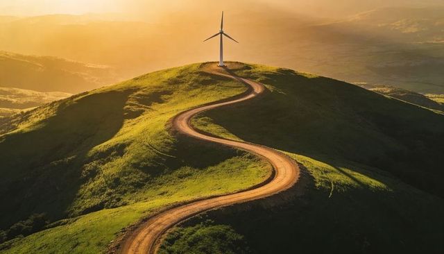S-shaped dirt road winding up green hill to wind turbine at golden sunrise