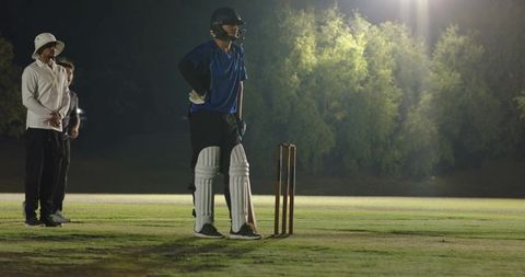 Night cricket batter standing at crease under stadium lights with on-field umpire and stumps