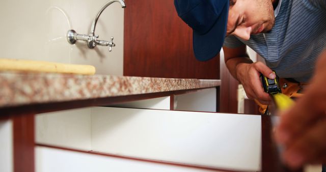 Handyman in blue cap measuring kitchen cabinets using measuring tape. Useful for illustrating home improvement tutorials, advertisements for renovation services, DIY project guides, and construction-related content.