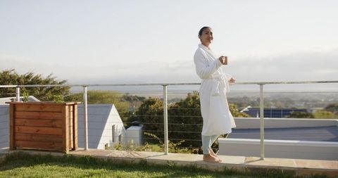 Mid-adult asian woman standing on balcony in white bathrobe holding coffee mug at sunrise
