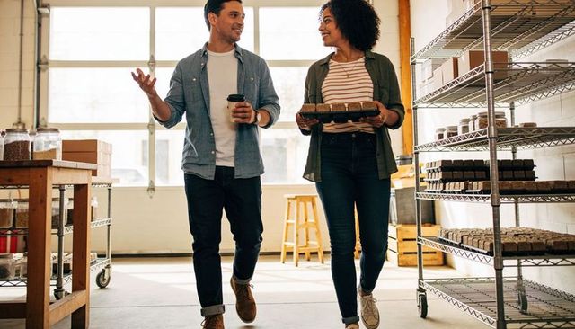 Small business owners walking through bakery workshop carrying muffin tray and coffee