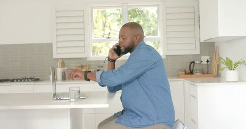 Black man working from home in bright modern kitchen talking on phone and pointing at laptop
