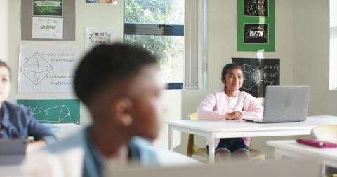 Elementary student working on laptop in bright classroom with math posters and sunlight