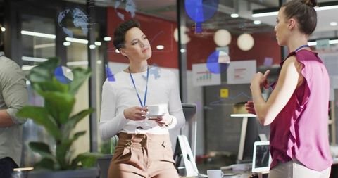 Woman holding coffee cup listening to colleague speaking by glass charts in modern office