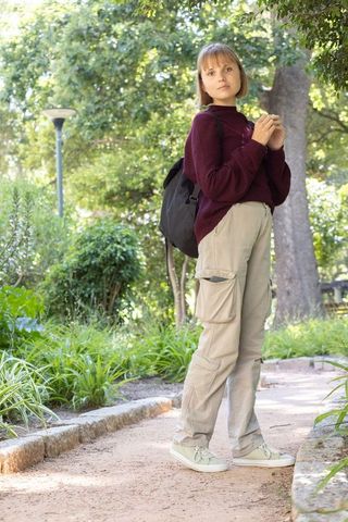 Young woman standing on sunlit park path wearing maroon sweater carrying black backpack