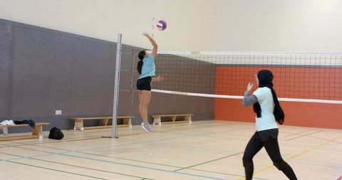 Diverse women jumping to block spike in indoor volleyball match featuring hijab player