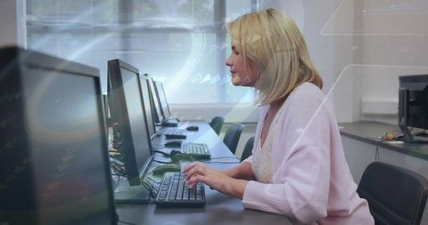 Mature woman typing on desktop in classroom wearing pink cardigan, education technology