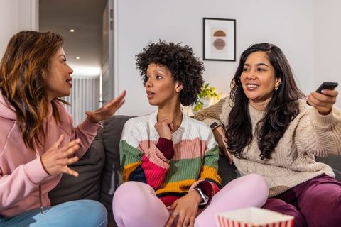 Three friends chatting on couch, watching movie and sharing popcorn in cozy living room