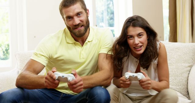 Happy couple sitting on sofa, playing video games together. Great for ads focusing on leisure activities, technology, family bonding, and home entertainment themes.