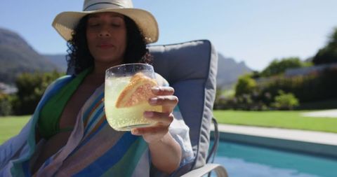 Poolside woman relaxing with citrus drink, straw hat and striped coverup at backyard pool