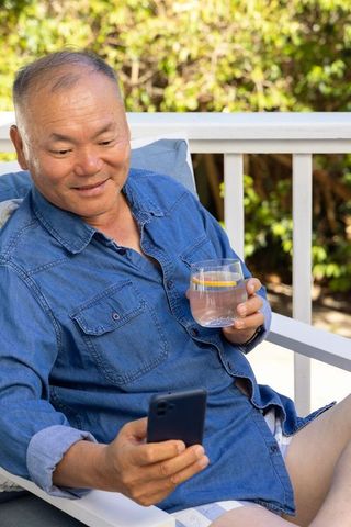 Senior asian man relaxing on porch holding lemon drink and smartphone, enjoying sunny deck