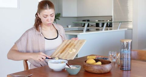 Young woman slicing banana into cereal, preparing healthy breakfast in modern minimalist kitchen