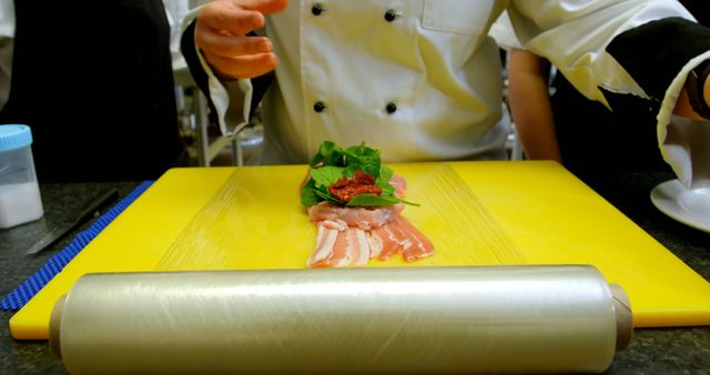 Chef arranging fresh greens and sun-dried tomatoes on slices of bacon on a yellow cutting board. Could be used in food blogs, culinary courses, or restaurant promotional materials showcasing food preparation processes.
