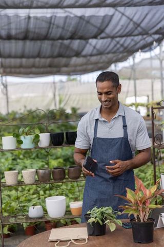 Indian man smiling and holding mobile while arranging potted plants in greenhouse nursery