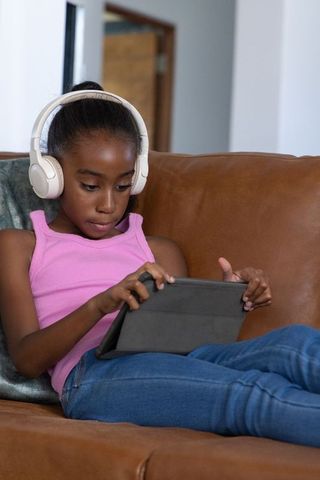 African american preteen using tablet with white headphones on brown couch for learning