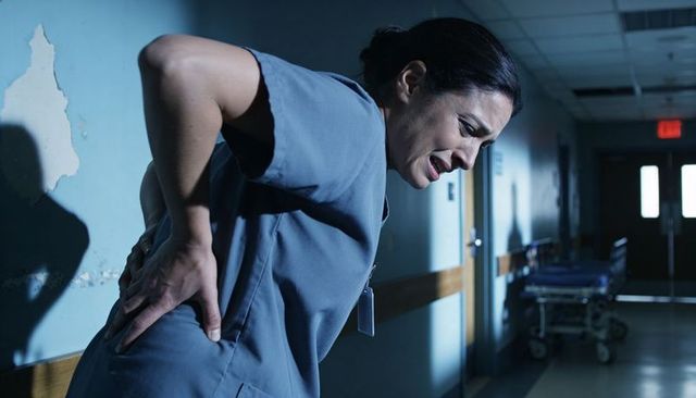 Night nurse clutching lower back in hospital corridor grimacing in pain near gurney