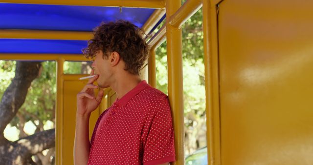 Young man in casual red polo shirt and jeans smoking a cigarette at a yellow bus stop shelter with a blue roof. He appears relaxed, enjoying his break in an urban setting on a sunny day. Ideal for use in campaigns related to smoking awareness, urban life, and youth culture.