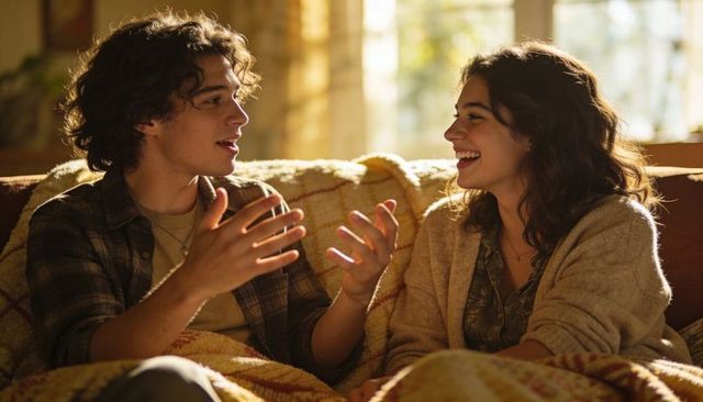 Sunlit cozy couple talking and laughing on sofa wrapped in warm blanket, intimate conversation