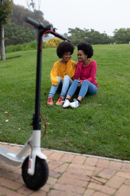 Front view of two happy biracial twin sisters enjoying free time together, embracing, smiling, sitting on grass in an urban park, electric scooter on a path in the foreground.