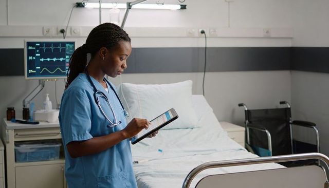 Nurse using tablet for clinical documentation by patient monitor in hospital room with wheelchair